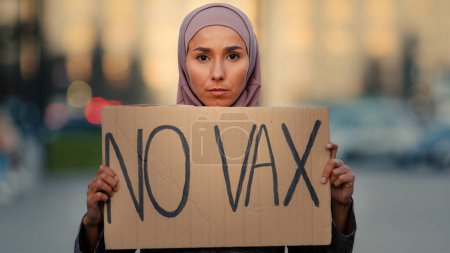 Photo for Portrait female muslim islamic girl woman in hijab standing in city outdoors showing cardboard banner sign text no vax protest against immunization covid19 coronavirus vaccination vaccine denial. High - Royalty Free Image