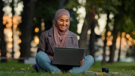 Photo for Happy smiling muslim islamic business woman girl female student wears hijab sitting on green grass lawn in city park using laptop for distant e-learning work chatting online checking mail with coffee - Royalty Free Image