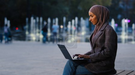 Téléchargez les photos : Jeune femme d'affaires musulmane réussie fille étudiante islamique porte hijab assis sur la rue ville en plein air arrière-plan urbain dactylographier ordinateur portable travailler en ligne e-learning à l'extérieur avec ordinateur chat app web - en image libre de droit