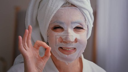 Photo for Close up old senior mature Caucasian woman with skincare textile skin mask on female face showing ok good beauty cosmetology procedure 60s lady wrap towel on head put moisturizing lifting cosmetic - Royalty Free Image