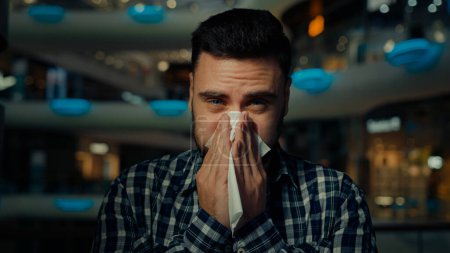 Foto de Hombre árabe enfermo joven hombre alérgico en el centro comercial sufren de secreción nasal gripe enfermedad alergia estacional cubierta boca estornudo limpiar la nariz con servilleta de papel tiene síntomas virales coronavirus infección - Imagen libre de derechos