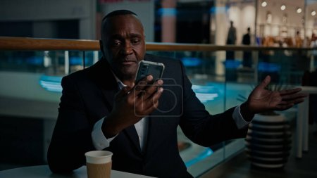 Photo for Ethnic African American man employer entrepreneur businessman in cafe with coffee tea talking business video call with mobile phone talk conference middle-aged African serious CEO shrug speak chatting - Royalty Free Image