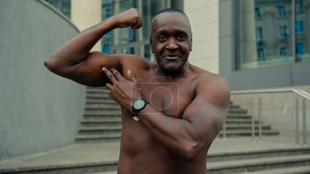 Photo for Middle-aged adult African American ethnic strong naked male athlete man bodybuilder showing biceps body muscles at web camera in city talk show fitness workout result talking hey you gesture outdoors - Royalty Free Image