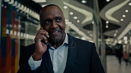 Foto de Sonriente alegre africano de mediana edad empresario hombre de negocios hablar de teléfono tiene negocio amigable negociación negociación en red con el teléfono inteligente en el interior explicando conversación llamada - Imagen libre de derechos