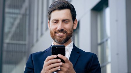 Photo for Smiling happy man holds phone browsing social network standing outdoors successful businessman responding to business or friendly email message male client makes order in online store using smartphone - Royalty Free Image