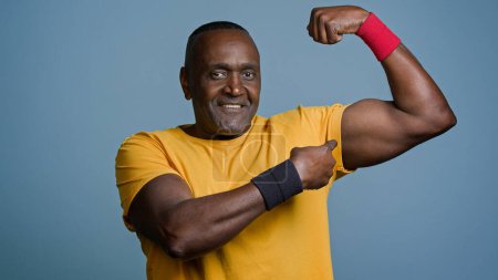 Photo for Smiling mature man sportsman standing in studio on gray background showing big manly muscles demonstrates pumped biceps athletic male trainer fitness instructor shows triceps motivates to play sports - Royalty Free Image