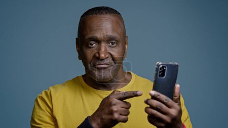 Photo for Mature man standing in studio on gray background holding mobile phone browsing social network sports athlete watching on internet physical exercise pointing to smartphone screen nods head approves - Royalty Free Image