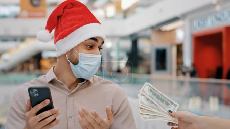 Photo for Ethnic man guy male in medical mask in X-mas Christmas Santa hat betting with mobile phone casino bet online bank payment credit service quick money cash prize reward financial winning loan winning - Royalty Free Image