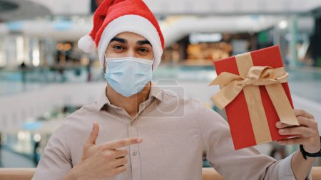 Photo for Man in Santa Christmas X-mas hat guy male in Covid face mask standing in shopping center mall looking camera showing gift present box wrapped in red paper celebrating New Year holiday in coronavirus - Royalty Free Image