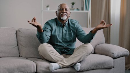 Téléchargez les photos : Happy elderly african american man sit on sofa in lotus position with folded fingers grandpa is meditating finds harmony relieves stress positive calm pensioner relaxing at home improve self-conscious - en image libre de droit