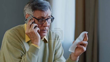 Photo for Old Caucasian elderly senior mature man grandfather male householder with check paper account bills finance talking mobile phone call to bank has problem with banking talk with financial consultant - Royalty Free Image