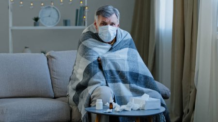 Photo for Ill sick old male in medical mask sitting on couch at home Caucasian elderly man wrapped in blanket feel unwell illness mature senior looking at camera covid-19 epidemic coronavirus outbreak flu. High - Royalty Free Image