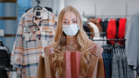 Photo for Blonde girl with long hair client buyer caucasian woman in medical mask in clothing store with packages looks into shopping bag surprised rejoices with purchases happy dances enjoying discounts sale - Royalty Free Image
