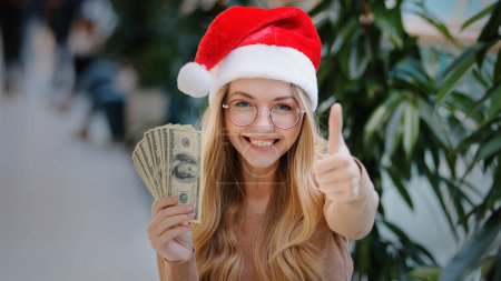 Photo for Happy rich successful Caucasian woman in red Santa Christmas hat cap and eyeglass holding money dollars counting cash prize success before shopping present financial discount to New year show thumb up - Royalty Free Image