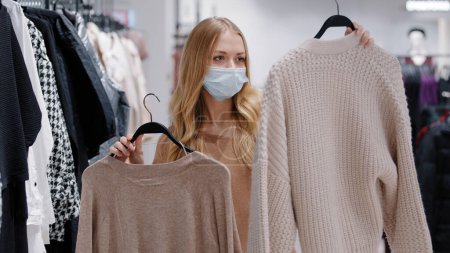 Photo for Young girl in medical mask choose clothes in store woman consumer make choice between two sweaters collects new fashionable image choosing jumpers thinking doubts lady shopping during pandemic covid19 - Royalty Free Image