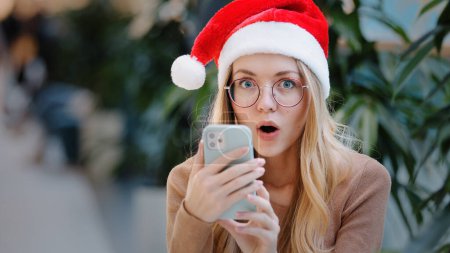 Photo for Close up funny happy shocked amazed Caucasian woman girl lady wearing Santa Claus Christmas hat and eyeglass looking at phone mobile win victory offer achievement New Year celebration winning discount - Royalty Free Image