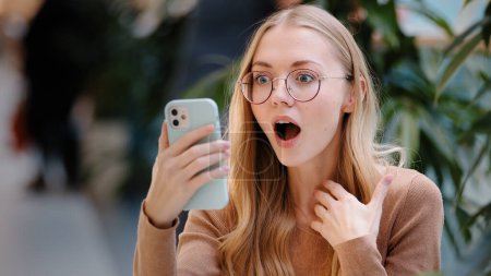 Foto de Portrait caucasian girl blonde successful lucky woman in glasses looks at screen of mobile phone wins online message with offer invitation happiness victory shock emotion good news with smartphone - Imagen libre de derechos