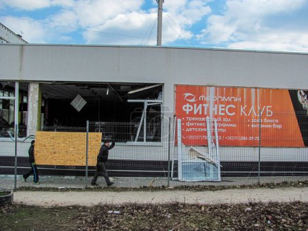 Foto de Kharkiv, Ucrania - 05.28.2022: destruido gimnasio de fitness quemado en Kharkov Ucrania hombres ucranianos de vidrio roto llevan fragmento de madera contrachapada de la puerta de la construcción después de la invasión rusa bombardeo guerra bombardeo arruinado ciudad - Imagen libre de derechos