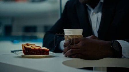 Téléchargez les photos : Gros plan recadré vue méconnaissable visiteur de café africain homme inconnu homme d'affaires en costume formel à la table à la cafétéria avec des brassages de gâteau mélanger le sucre dans une tasse de café thé latte cappuccino avec bâton de bois - en image libre de droit