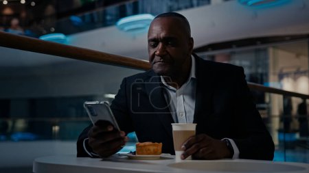 Foto de Africano adulto de mediana edad empresario de 50 años hombre étnico en traje formal empresario sentado a la mesa en la cafetería beber café beber té navegación teléfono móvil teléfono inteligente charla decir videollamada - Imagen libre de derechos