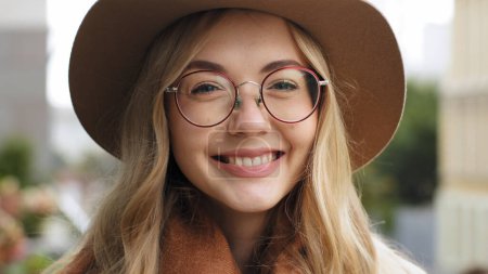 Téléchargez les photos : Gros plan souriant Caucasien 20s femme sourire largement avec des dents blanches en regardant la caméra fille heureuse portant des lunettes et chapeau posant dans la rue en ville dame millénaire avec maquillage naturel à l'extérieur - en image libre de droit