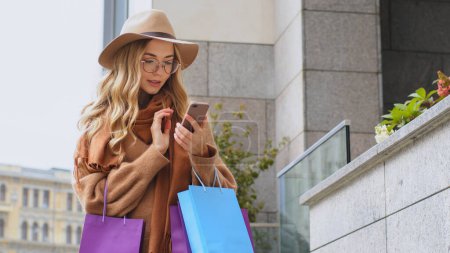 Photo for Stylish fashionable buyer client consumer female Caucasian girl woman with shopping bags purchases outdoors in city near mall browsing mobile phone buying ordering online payment in Internet shop app - Royalty Free Image