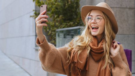 Photo for Laughing smiling female shopper buyer client girl talking video call make photo with mobile phone Caucasian woman on street with shopping bags lady with hat and glasses outdoors talk conference chat - Royalty Free Image