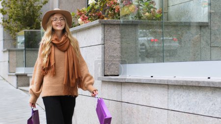 Téléchargez les photos : Fille caucasienne en chapeau à la mode marchant sur la rue après avoir fait du shopping avec des paquets 20s femme consommateur client acheteur femme dame souriante marcher à l'extérieur avec des sacs en plein air profiter de rabais de vente Black Friday - en image libre de droit