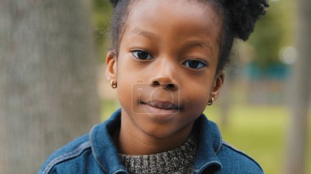 Téléchargez les photos : Petite fille afro-américaine en plein air gros plan enfant joli sourire en regardant la caméra écolière ethnique debout dans le parc portrait mignon petit élève enfant fille visage en ville headshot garde. Haut - en image libre de droit