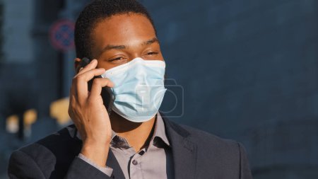 Téléchargez les photos : African American businessman ethnic man male guy in medical face mask talking mobile phone answer call talk with smartphone calling speaking with cellphone in city outdoors coronavirus covid pandemic - en image libre de droit
