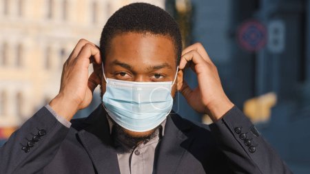 Téléchargez les photos : Headshot portrait in city outdoors African American businessman put on protective medical face mask looking at camera. Ethnic man male guy entrepreneur in respiratory protect coronavirus outbreak - en image libre de droit