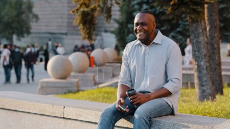 Foto de Happy smiling African American ethnic middle-aged man businessman male sitting outdoors in city with coffee tea and phone waiting for date business meeting smile to friend carefree cheerful relaxing - Imagen libre de derechos