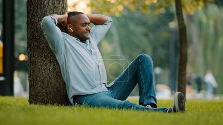 Téléchargez les photos : Calm relaxed resting middle-aged male listening music with earphones sitting on green grass in park under tree enjoy song relaxing adult African American ethnic man in wireless headphones listen audio - en image libre de droit