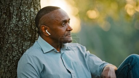 Photo for Headshot portrait close up elderly middle-aged adult African American man businessman relaxing sitting under tree in city park work break listening music audio song sound with wireless headphones - Royalty Free Image