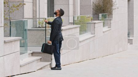 Foto de African American ethnic man candidate to job offer work interview standing near office building looking at wristwatch. Businessman entrepreneur hurry to business meeting check time standing in city - Imagen libre de derechos