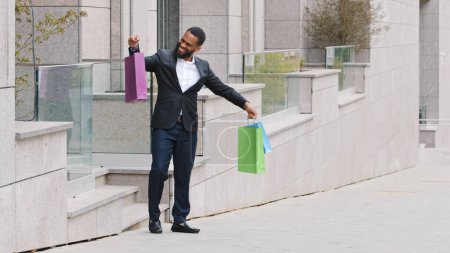 Téléchargez les photos : Millennial male ethnic customer client buyer businessman African American man walking out of mall boutique store walk in city with shopping bags happy holding purchases clothes enjoy discounts offer - en image libre de droit