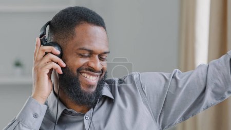 Téléchargez les photos : Happy African American man office worker ethnic businessman manager entrepreneur executive in headphones listening to music sitting at workplace funny relaxed guy enjoy favorite audio melody dancing - en image libre de droit