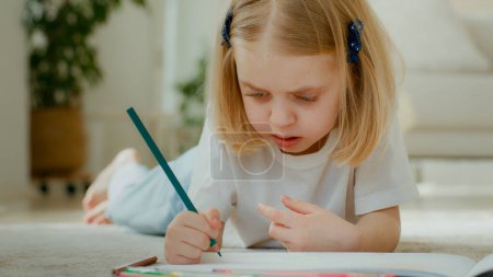 Photo for Cute concentrated Caucasian little kid girl draw color pencil lying on floor at home play alone. Talented preschool child daughter kid baby coloring picture at home drawing painting creative paint - Royalty Free Image