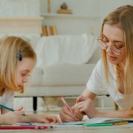 Little small adopted child girl enjoy art draw with Caucasian mother in glasses lying at home floor together. Daughter baby kid with mommy mom babysitter teacher drawing picture with pencils talking