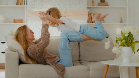 Photo for Caucasian mother in glasses strong mom lying on cozy couch on back holding little daughter in air on legs bent in knees play airplane flying game child girl preschooler kid baby imitate flight plane - Royalty Free Image