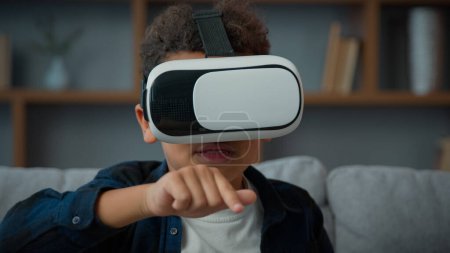 Foto de Little African American ethnic boy pupil son kid child schoolboy at home wearing VR glasses play online video 3d game education studying with augmented virtual reality headset AR technology playing - Imagen libre de derechos