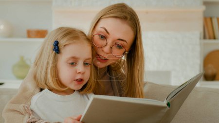 Téléchargez les photos : Caucasian mother in eyeglasses with little small cute daughter preschool child girl baby reading book in living room at home mom with kid read literature at couch sofa elementary learning education - en image libre de droit
