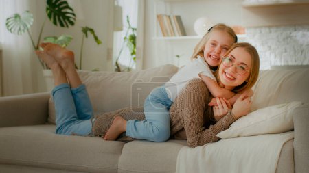 Téléchargez les photos : Caucasian mother in glasses lying on couch sofa in home living room little cute daughter kid child baby girl embrace hug cuddle mom by neck piggyback laughing together relaxing enjoy hugging cuddling - en image libre de droit