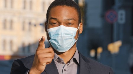 Téléchargez les photos : African American businessman ethnic man male guy in medical face mask pointing with finger hey you agree show number one support gesture choosing choice in city outdoor recommend pandemic covid safety - en image libre de droit