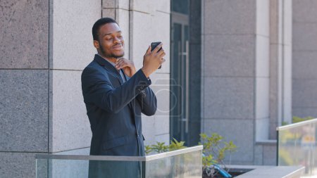 Foto de African American office worker standing on balcony speaking with colleagues with smartphone video chat app. Ethnic businessman entrepreneur manager employee has mobile phone distant call conferencev - Imagen libre de derechos