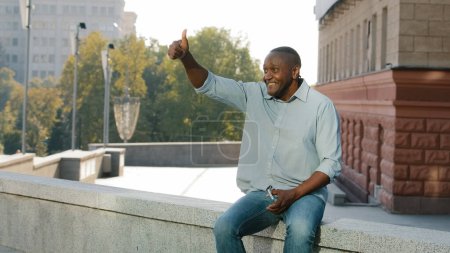 Foto de Friendly middle-aged adult mature African American man businessman entrepreneur male sitting outdoors in city waiting for meeting date waving hand welcoming friend colleague invite come here calling - Imagen libre de derechos