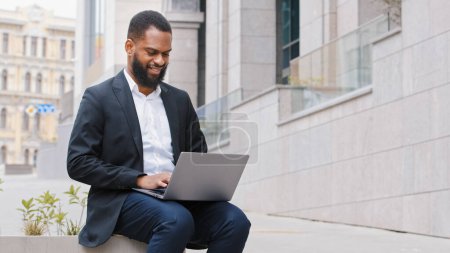 Photo for Male African American millennial freelancer worker entrepreneur employee businessman ethnic man typing on laptop outdoors bearded professional work with online computer technology app sitting in city - Royalty Free Image