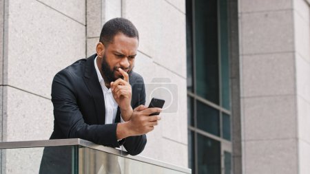 Téléchargez les photos : Pensive thoughtful ethnic African American bearded man looking at smartphone screen think about business idea pondering browsing cellphone thinking businessman with mobile phone standing on balcony - en image libre de droit