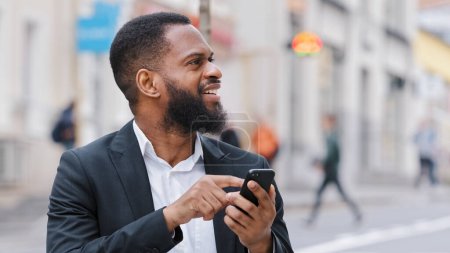 Foto de Smiling ethnic African American bearded man businessman executive employer with smartphone in city chatting shopping online looking away booking taxi in mobile app using application GPS map in phone - Imagen libre de derechos