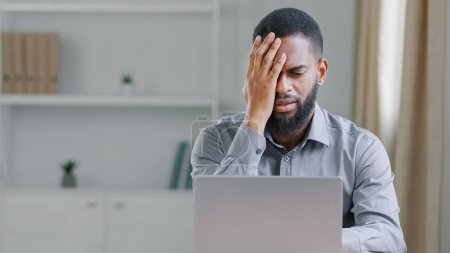 Foto de Stressed disappointed African American ethnic bearded man holding head with headache tired overworked manager reading bad news office worker businessman with laptop computer failure online problem - Imagen libre de derechos